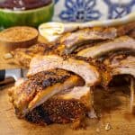 Braised pork ribs sliced and arranged on a wooden cutting board, accompanied by bowls of seasoning and BBQ sauce, set against a blue and white tile background.