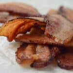 A close-up shot of perfectly crispy bacon slices stacked on top of each other, showcasing their rich brown color and slightly curled edges, placed on a white parchment paper background.