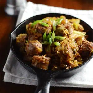 A cast iron skillet with bratwurst and cabbage, garnished with green onions, placed on a white cloth napkin on a wooden table.