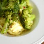 Close-up of steamed broccoli florets seasoned with pepper flakes, served alongside creamy scrambled eggs on a white plate.
