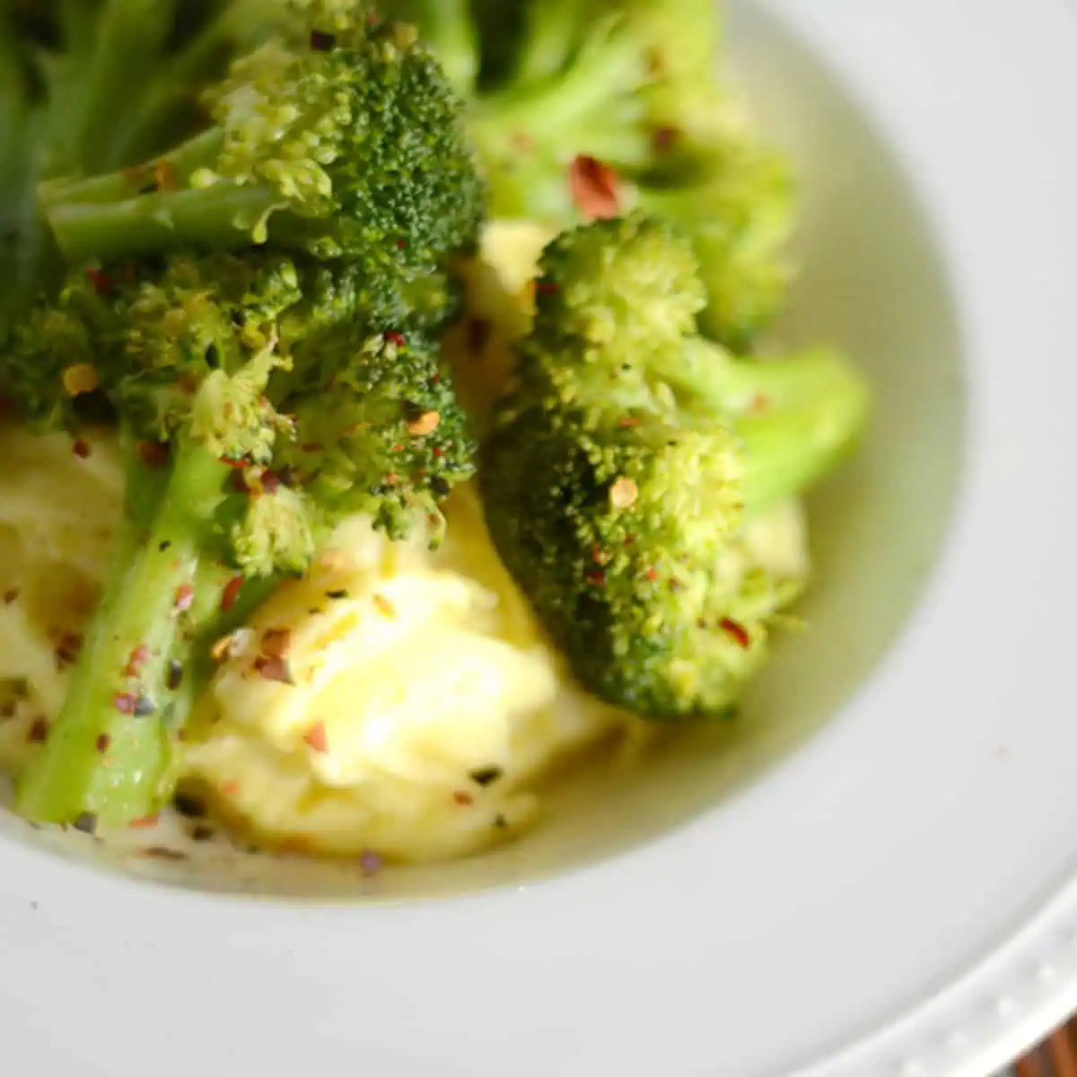 Close-up of steamed broccoli florets seasoned with pepper flakes, served alongside creamy scrambled eggs on a white plate.