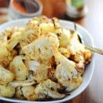 Roasted cauliflower florets served in a white bowl, with a background of spices and seasoning jars.