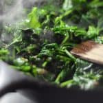 A close-up of fresh spinach being sautéed in a skillet with steam rising, showing a wooden spatula stirring the vibrant green leaves.