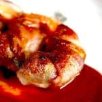 Close-up of bacon-wrapped shrimp with a side of vibrant red sauce, highlighting the crispy texture and rich color of the bacon.