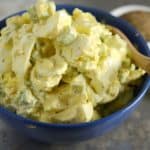 A blue bowl filled with creamy egg salad, featuring chunks of boiled eggs and diced celery, garnished with a sprinkle of pepper. A small dish of pepper and a stalk of celery are in the background.