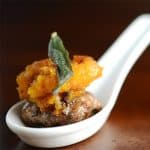A small white spoon holding an appetizer of butternut squash atop a sausage patty, garnished with a crisp sage leaf.