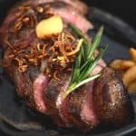 Close-up shot of a sliced pan-seared flat iron steak in a cast iron skillet, topped with crispy shallots, a garlic clove, and a sprig of rosemary.