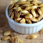 A white ramekin filled with roasted pumpkin seeds, some of which have spilled onto the textured surface around it, showcasing their golden-brown, crispy texture.