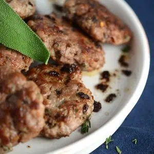 A plate of homemade sage sausage patties with a fresh sage leaf garnish, shown on a blue tablecloth, highlighting the texture and herbs in the sausage.