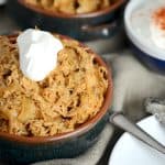 Close-up of a bowl of shredded chicken paprikash topped with sour cream. The background includes another bowl of the dish and a small bowl of sour cream with paprika, set on a textured cloth.