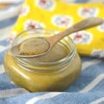 A small glass jar filled with honey mustard sauce, with a wooden spoon resting on top, holding a spoonful of the sauce. The jar is placed on a blue and white striped cloth, with a yellow floral napkin in the background.