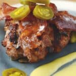 Grilled pork burger topped with crispy bacon, jalapeño slices, and drizzled with creamy yellow sauce, served with pickles on a gray plate.