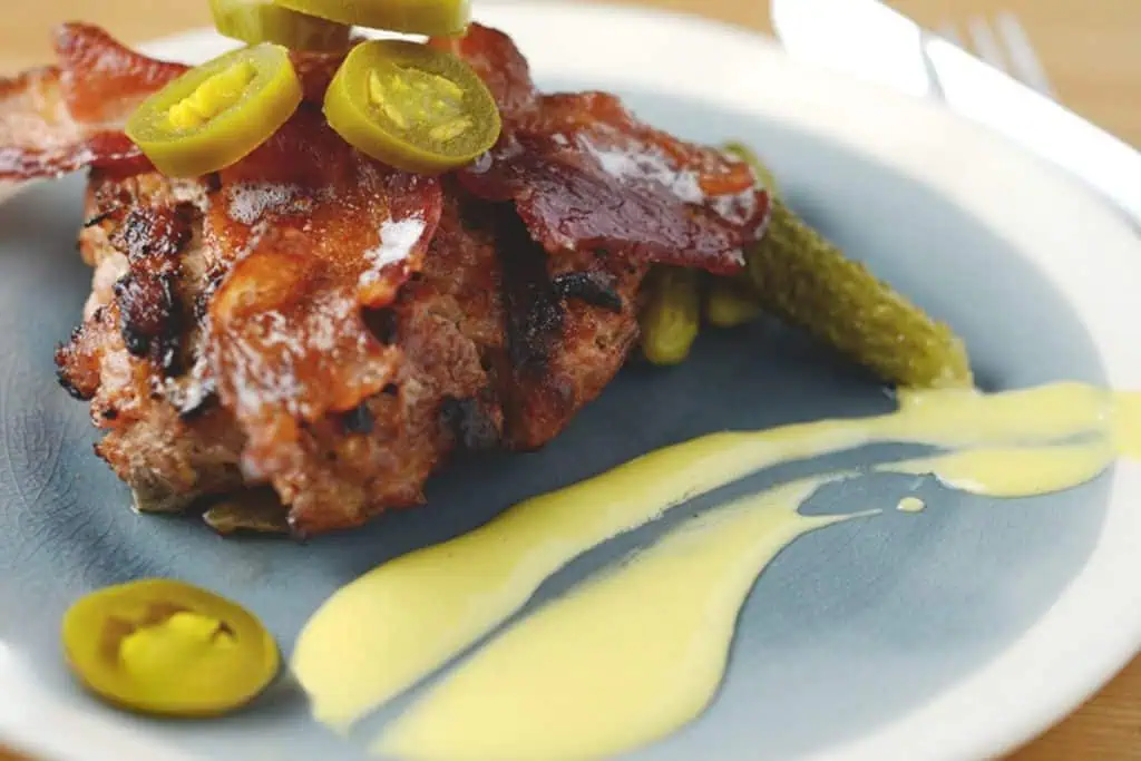 Grilled pork burger topped with crispy bacon, jalapeño slices, and drizzled with creamy yellow sauce, served with pickles on a gray plate.
