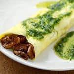A savory omelette filled with wild mushrooms, topped with a green herb sauce, served on a white plate.