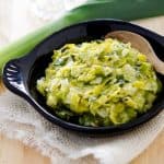 Close-up of a black dish containing creamy, seasoned leeks, with a wooden spoon for serving and fresh leeks in the background.
