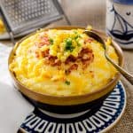 A hearty bowl of cheesy grits, topped with bacon, corn, and parsley, placed on a blue and white plate. A grater is visible in the background.