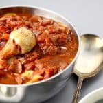 Close-up of a bowl of marinara sauce with visible chunks of tomatoes, onions, and an artichoke heart, with a spoon resting beside it.