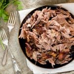 A plate of easy pulled pork served on a dark plate with two forks beside it, placed on a linen cloth, with a bunch of cilantro in the background.