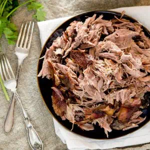 A plate of easy pulled pork served on a dark plate with two forks beside it, placed on a linen cloth, with a bunch of cilantro in the background.