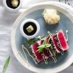 Close-up of seared ahi tuna slices with a black and white sesame seed crust, garnished with green onions, served on a blue plate with wasabi and soy sauce in small bowls on the side.