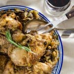 A detailed shot of a blue plate filled with coq au vin, highlighting the juicy chicken pieces and mushrooms, garnished with a fresh rosemary sprig, with a glass of red wine in the background.