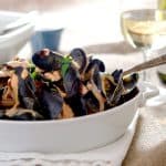 A bowl of freshly cooked mussels in a creamy, tangy sauce, garnished with fresh herbs, with a glass of white wine in the background.