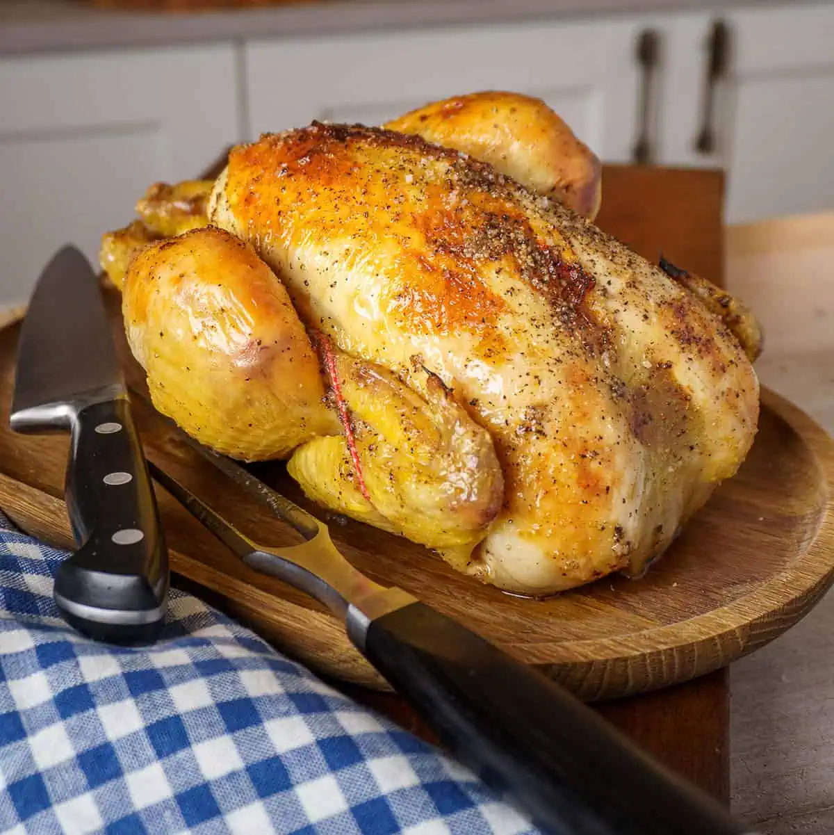 Side view of a golden-brown roasted chicken on a wooden platter, ready to be carved. A carving knife and fork are set beside the platter, with a blue and white checkered cloth in the background.