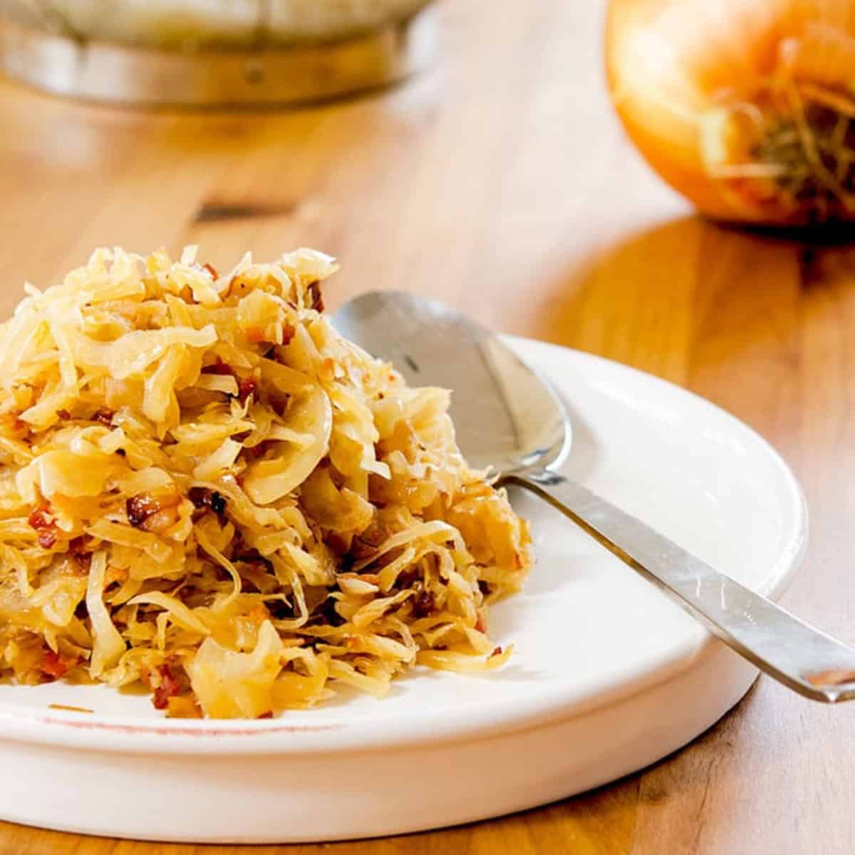 A serving of sauerkraut on a white plate with a spoon, placed on a wooden table. In the background, there is a whole onion and a colander with shredded cabbage.