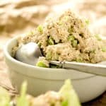 A bowl of low-carb tuna salad with diced celery and onions, placed on a textured surface with a spoon inside the bowl.