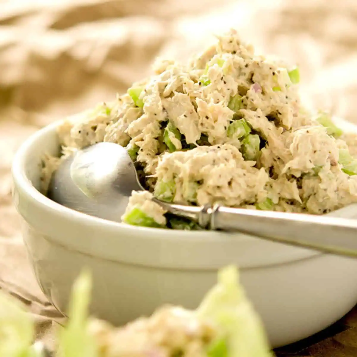 A bowl of low-carb tuna salad with diced celery and onions, placed on a textured surface with a spoon inside the bowl.