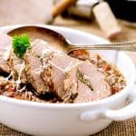 Sliced roasted pork tenderloin served in a white dish, topped with fresh parsley and accompanied by a savory vegetable sauce, showcasing a rich and hearty presentation.