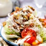 Close-up of a loaded iceberg wedge salad topped with creamy dressing, crumbled bacon, blue cheese, walnuts, and cherry tomatoes.