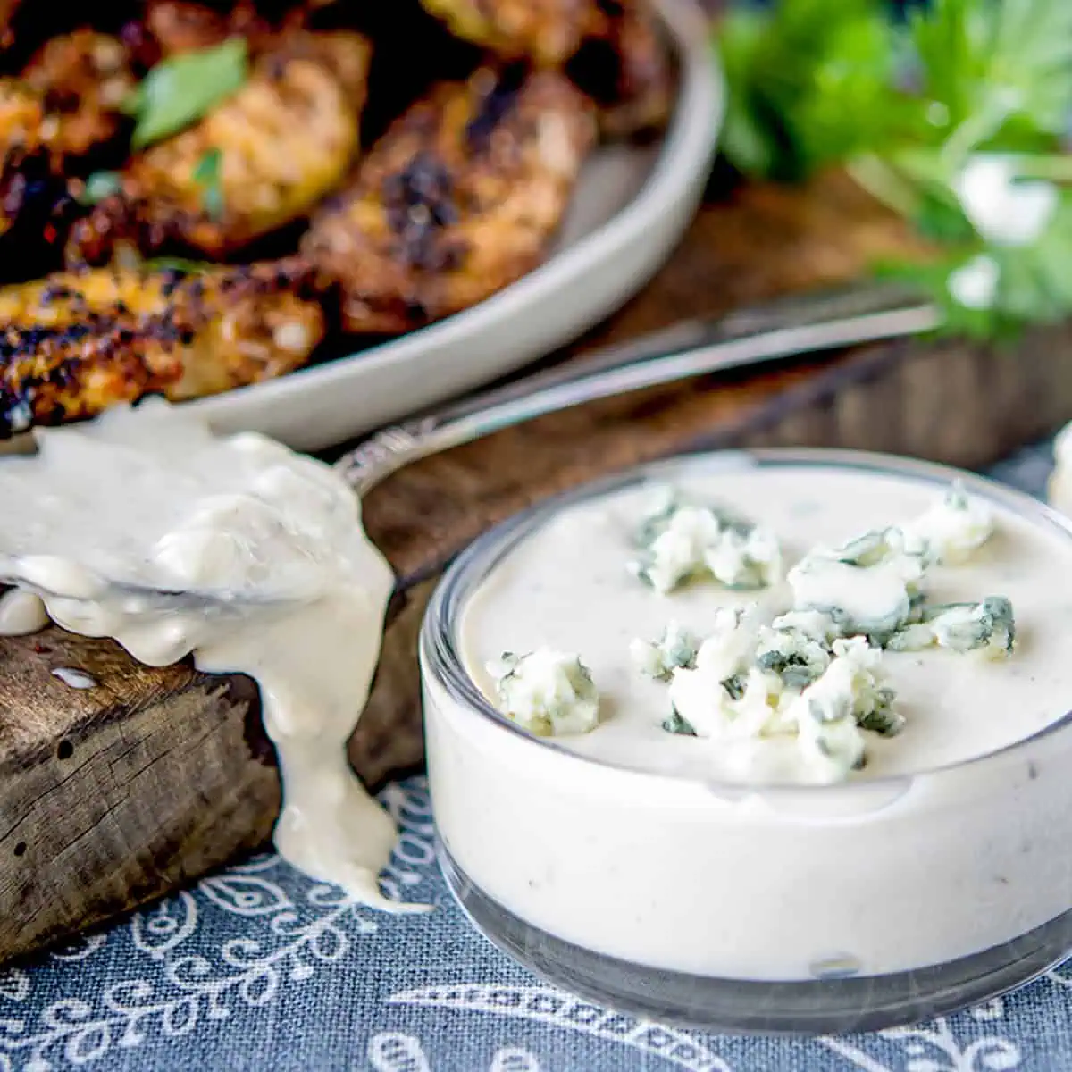 A close-up view of a spoon dripping creamy blue cheese dressing beside a small bowl filled with the same dressing topped with crumbled blue cheese. In the background, there are grilled chicken tenders on a plate.