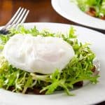 A close-up of a plate of frisée salad topped with a perfectly poached egg, with stacked white plates and cutlery in the background.
