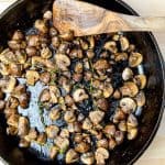 An overhead view of roasted garlic sautéed mushrooms in a cast-iron skillet, showing the even distribution of mushrooms and thyme sprigs, with a wooden spatula.