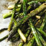 A close-up of roasted asparagus spears on a baking sheet, glistening with olive oil and sprinkled with cracked black pepper and kosher salt.