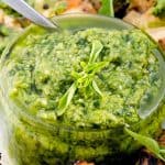 A close-up of a glass jar filled with vibrant green basil pesto, garnished with a sprig of fresh basil, with a silver spoon resting on the side.