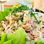 Close-up of rotisserie chicken salad garnished with chives and served on a bed of green lettuce. A single chive is placed as a decorative touch on top. The background shows a glass of rosé wine, suggesting a light and refreshing meal setting.