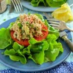 Fresh tuna salad served on a thick slice of tomato, garnished with chopped chives, and surrounded by crisp green lettuce on a blue plate. A vintage fork and a wedge of lemon are placed beside the plate.
