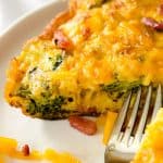 Close-up of a slice of bacon and egg frittata on a white plate, showing the golden, crispy edges, vibrant green broccoli, and melted cheddar cheese with bits of bacon.