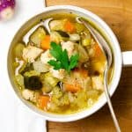 A centered, overhead view of a bowl of chicken vegetable soup on a wooden surface. The soup features a variety of colorful vegetables and chicken pieces, garnished with a fresh parsley leaf, with a silver spoon placed in the bowl.