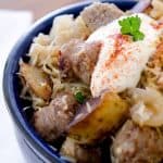 A close-up view of sauerkraut and sausage pieces in a blue bowl, garnished with sour cream and paprika, with a fork and napkin on the side.