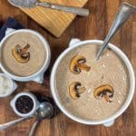 Two bowls of creamy mushroom soup garnished with sautéed mushroom slices, set on a wooden table with a spoon and peppercorns in the background.