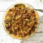 A beautifully browned Brussels sprouts gratin in a glass baking dish, topped with a golden, crispy crumble. The gratin is garnished with finely chopped parsley, adding a touch of green to the dish.