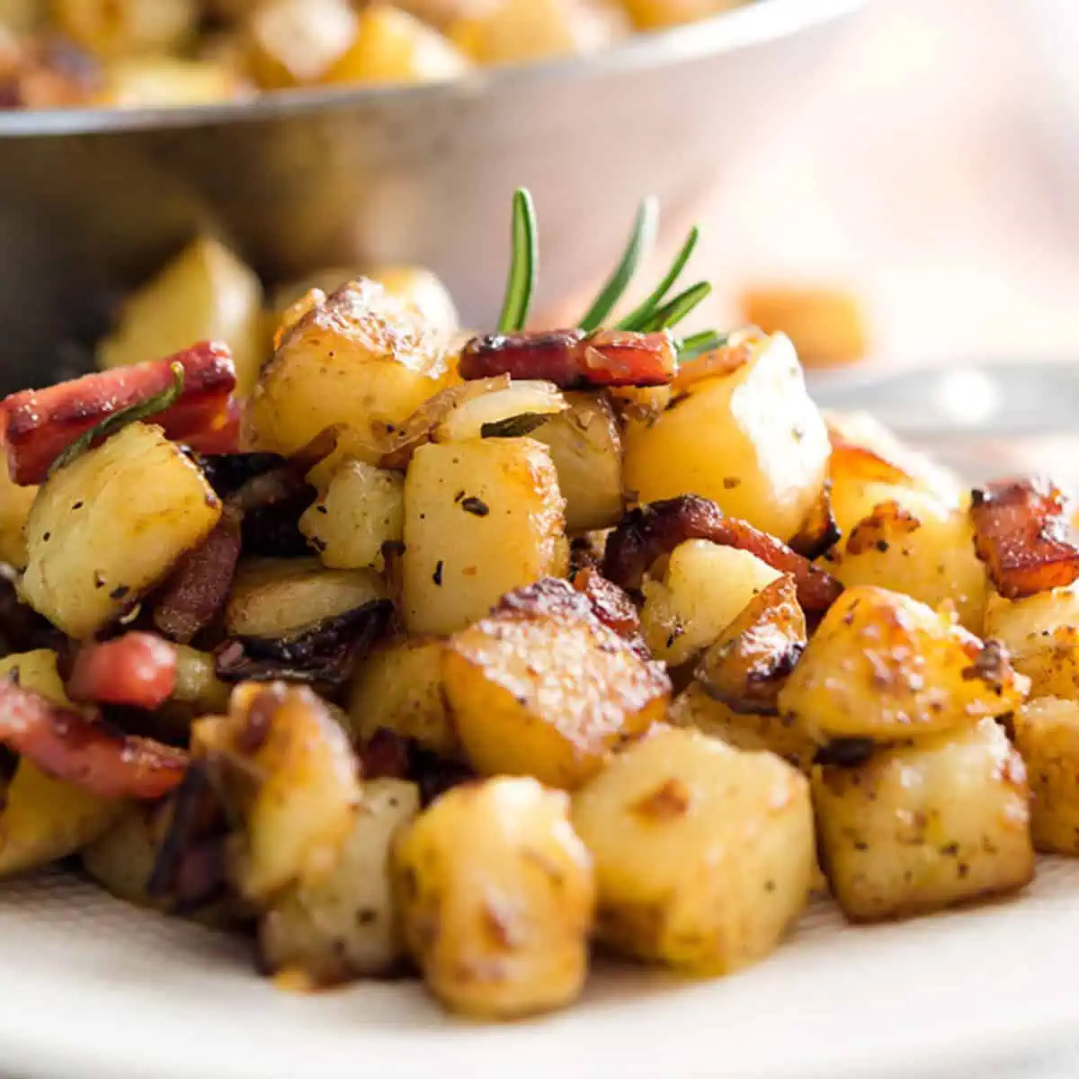A close-up of a plate of homemade hash browns with crispy bacon pieces, garnished with rosemary, in front of a skillet of more hash browns.