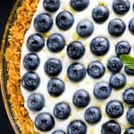 Overhead view of a lemon no-bake cheesecake garnished with fresh blueberries and lemon zest, set on a clear pie dish with a golden crust.