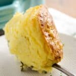 A spoon lifting a serving of fluffy, golden-brown topped cream cheese mashed potatoes from a turquoise baking dish.