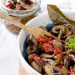 Marinated mushrooms with red bell peppers and a bay leaf in a bowl with a wooden spoon, with a glass jar of the same marinated mushrooms in the background.