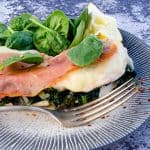 Close-up view of a Chicken Saltimbocca dish on a decorative plate. The dish features a tender chicken breast topped with melted cheese, a slice of prosciutto, and fresh spinach leaves, garnished with a wedge of lemon and red pepper flakes. A fork rests beside the chicken, ready for serving.