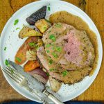 A plate with slices of leg of lamb drizzled with gravy, accompanied by roasted root vegetables and garnished with chopped parsley.